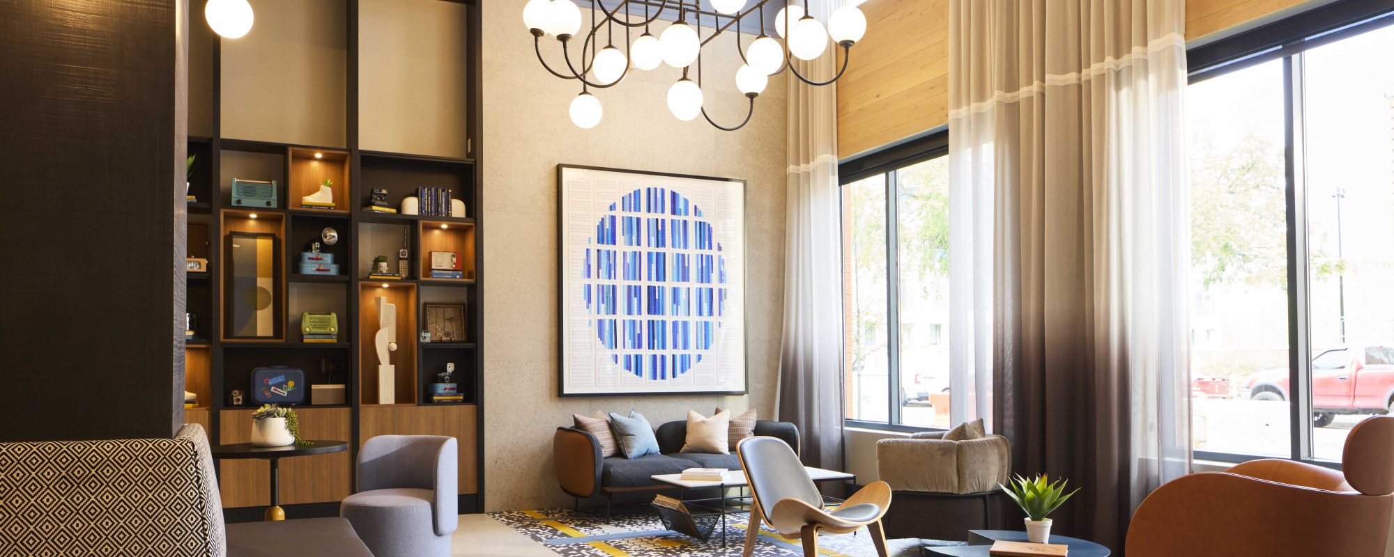 Modern lounge with large windows, contemporary furniture, a geometric blue artwork on the wall, shelves with decor items, and a chandelier hanging from the ceiling.