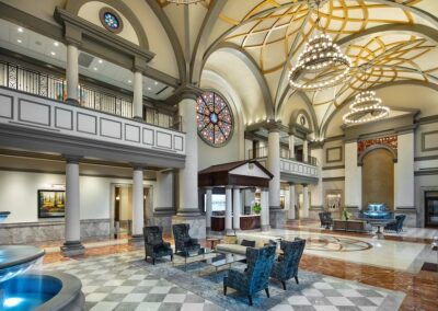 Spacious, elegant hotel lobby with high arched ceilings, chandeliers, a stained glass window, marble floors, seating area, and a fountain.