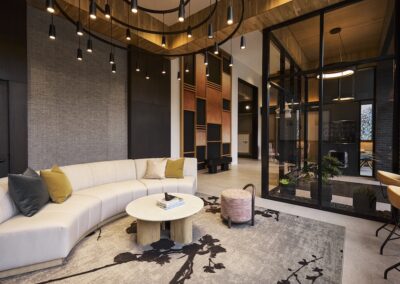 Modern lounge with a curved beige sofa, round coffee table, decorative rug, hanging lights, and a glass-walled room with plants in the background.