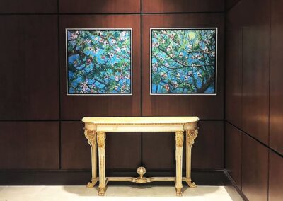 A cream and gold ornate console table stands against a dark wood-paneled wall, with two framed paintings of blossoming tree branches hanging above it.
