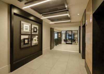 Modern hallway with framed pictures on a dark accent wall, light tile flooring, recessed ceiling lights, and glass doors leading to an office area in the background.