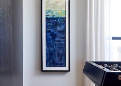Tall abstract painting with blue and white tones hangs on a white wall beside a foosball table in a modern room with wood flooring and sheer curtains.