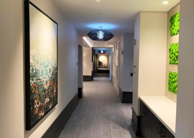 A modern hallway with tile flooring, wall art, ceiling lights, and a section of green plant decor on the right wall.