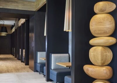 Modern restaurant interior featuring blue upholstered booth seating, wooden tables, black paneled walls, and a tall wooden sculpture of stacked, smooth, rounded forms.