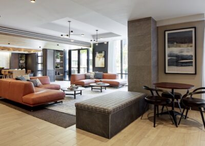 Modern lobby with orange sofas, a checkered bench, round table with chairs, large windows, wood flooring, and wall art. Natural light filters into the space.