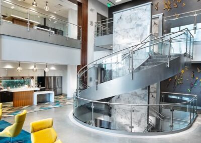 Modern office lobby with yellow chairs, a spiral glass staircase, marble wall accents, and a kitchen area in the background. Sculpted birds decorate one wall.