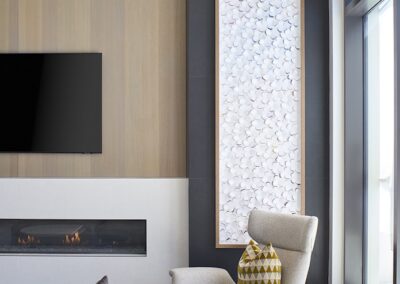 Modern living room with a wall-mounted TV, linear fireplace, beige armchair, geometric pillows, large artwork, and floor-to-ceiling windows.