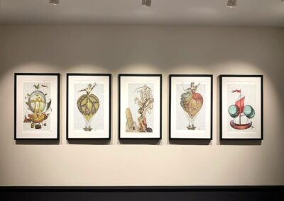 Five framed art prints featuring whimsical vintage-style illustrations of ships and hot air balloons are displayed in a row on a white wall under spotlights.