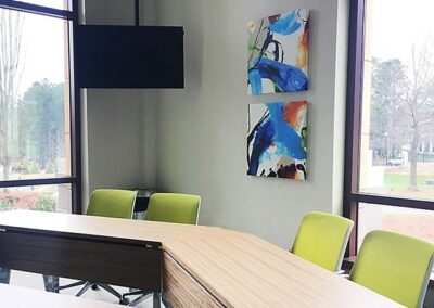 Modern conference room with light wood tables arranged in a U-shape, green chairs, a wall-mounted TV, abstract art, and large windows showing an outdoor view.