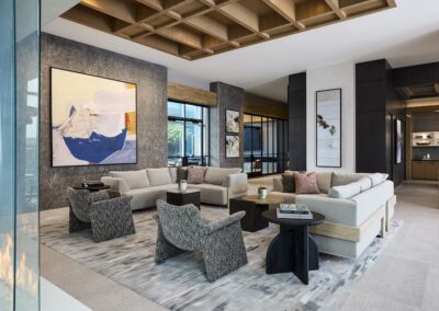 Modern living room with light-colored sectional sofas, patterned armchairs, abstract wall art, and a wooden ceiling accent, featuring large windows and a neutral color palette.