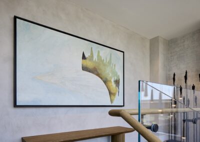A framed abstract painting with muted colors hangs on a light gray wall above a wooden console table and beside a glass stair railing.