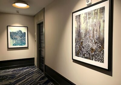 A hallway with two framed abstract artworks on cream-colored walls, a closed dark door, patterned carpet, and a ceiling light fixture.