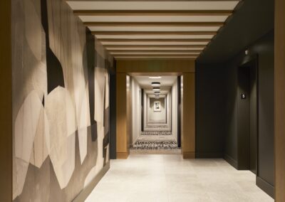 A modern indoor hallway with a patterned carpet, abstract wall art on the left, and elevator doors on the right, leading to a vanishing point.