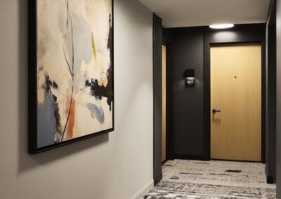 A modern hallway with abstract artwork on the left wall, patterned carpet, and a closed wooden door labeled 107 at the end. Ceiling lights illuminate the space.