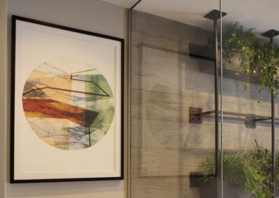 Framed abstract artwork with geometric shapes hangs on a beige wall next to a glass partition with green plants on shelves.