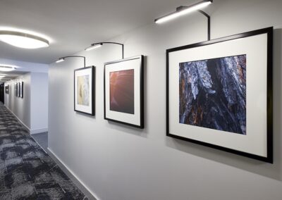 A hallway with patterned carpet and framed abstract photographs on the wall, each illuminated by individual picture lights.