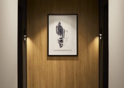 A framed abstract black and white artwork hangs on a wooden wall in a hallway with patterned carpet and ceiling light.
