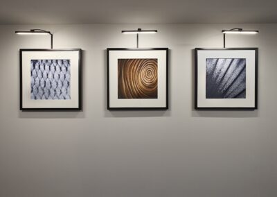 Three framed abstract art pieces are mounted on a wall, each illuminated by an overhead picture light.