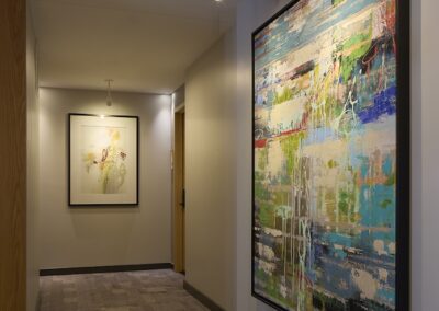 A hallway with gray tile flooring features large abstract paintings on the walls, illuminated by overhead spotlights.