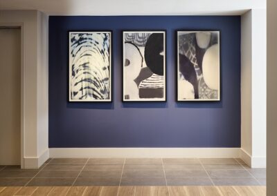 Three abstract, black-and-white framed artworks hang on a blue accent wall in a modern hallway with tiled and wooden flooring.
