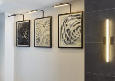 Three abstract black-and-white framed artworks are mounted on a light wall, each illuminated by individual picture lights above them. A modern vertical light fixture is on a dark wall to the right.