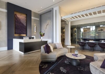Modern hotel lobby with a reception desk, curved seating, a round coffee table, decorative wall art, and a row of purple chairs near a glass wall.