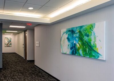 A hallway with light gray walls and two colorful abstract paintings, one on the side wall and another at the end of the hallway, under bright ceiling lights.