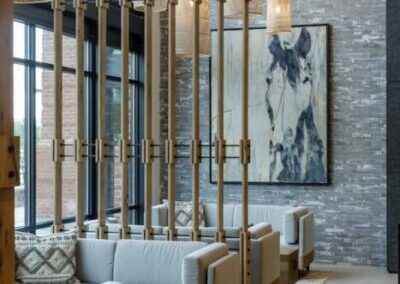 Modern lounge area with gray armchairs, wooden floor, hanging wicker lamps, a large window, and a horse painting on a gray brick wall.