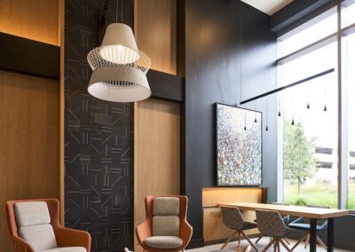 Modern lobby with tall windows, orange and gray chairs, a round coffee table, pendant lighting, wall art, and a wooden accent wall. Natural light fills the space.