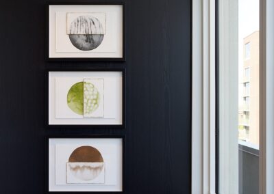 Three framed abstract artworks featuring circular shapes in black, green, and brown tones hang vertically on a dark wall next to a large window.