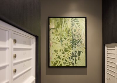 A framed abstract green and white painting hangs on a beige wall between rows of white mailboxes with numbered brass knobs.