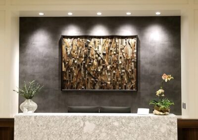 A marble reception desk with a vase of greenery and an orchid, set against a dark wall with a large abstract wooden art piece and overhead lighting.