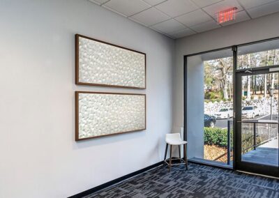 A modern office lobby with two abstract paintings on a white wall, a white chair in the corner, glass doors, and a view of parked cars and trees outside.
