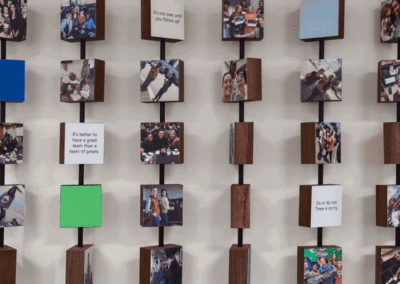 A wall display of wooden blocks featuring photos of people, blank colored squares, and motivational quotes, all mounted on vertical rods.