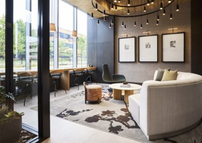 Modern office lounge with a white curved sofa, round coffee table, two chairs, wall art, pendant lights, and a row of desks by large windows overlooking greenery.