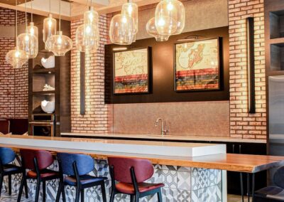 Modern bar area with hanging glass pendant lights, a patterned tile floor, a long counter with mixed-color chairs, brick walls, and framed art pieces above the sink.