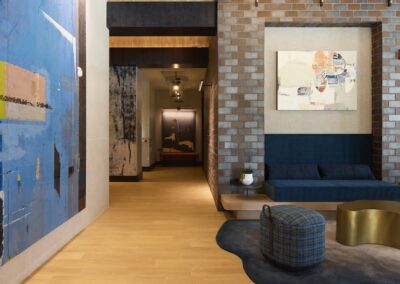 Modern lobby area with abstract artwork, a built-in blue bench, round plaid ottoman, gold accent table, and wood and brick wall elements.