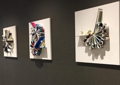 Five square white panels with rolled and folded magazine paper sculptures are mounted in a row on a dark wall, each displaying unique abstract designs.
