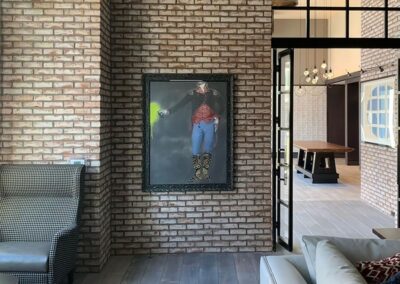 A brick wall with framed artwork of a cowboy holding a bright green object, next to a checkered chair and a beige sofa in a modern living space.