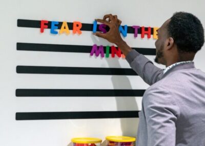 A man arranges colorful magnetic letters on a wall-mounted board; the phrase "FEAR IS IN THE MIND" is partially spelled out.