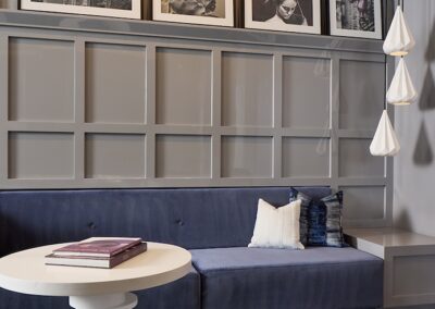 A modern lounge with a blue upholstered bench, white round table, small stool, three pendant lights, and framed art arranged on a gray paneled wall.