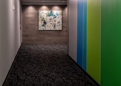 A hallway with patterned carpet, colorful green and blue panels on the right wall, and an abstract painting on the far end wall under overhead lighting.