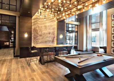 Modern lounge area with a pool table, bar seating, wood floors, large windows, decorative lighting, and framed wall art.