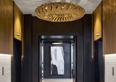 Elevator lobby with two black elevator doors, wood paneling, a decorative ceiling light fixture casting shadows, and a large abstract artwork at the end of the hallway.