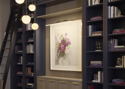 Modern library with navy built-in bookshelves, a rolling ladder, and a large floral artwork framed by shelves. Round pendant lights hang from the ceiling.