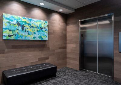 A modern elevator lobby with wood-paneled walls, a black bench, abstract artwork, and closed stainless steel elevator doors.