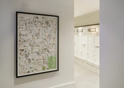 Framed abstract art with white and green squares hangs on a white wall next to a hallway with white mailboxes and wall lights.