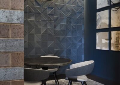 A modern meeting nook with a round black table, three gray chairs, geometric textured blue wall, abstract art, and a large circular ceiling light.