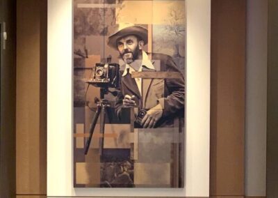 A sepia-toned portrait of a man with a hat holding a vintage camera, displayed on a wall in a modern, well-lit hallway under an abstract ceiling light.