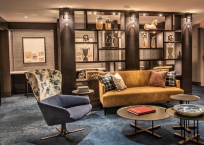 Modern lounge area with a mustard sofa, blue chair, nested coffee tables, and a backlit shelving unit displaying decorative items and books.